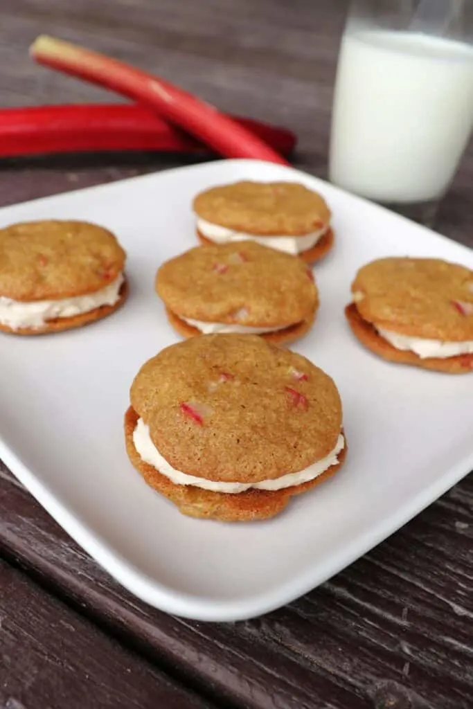 5 rhubarb cookie sandwiches with caramel filling on a square white plate. Fresh rhubarb stalks and a glass of milk sit in the background.