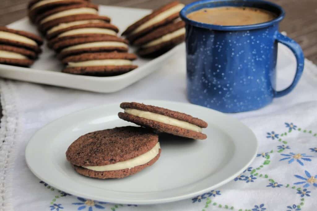 2 chocolate sandwich cookies sitting on a plate, with a platter of more cookies in the background and a blue tin cup full of coffee.