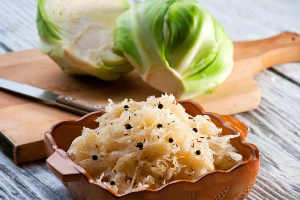 A bowl of sauerkraut sitting in front of a cutting board with a knife and fresh head of cabbage.