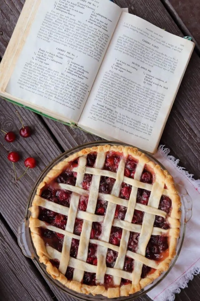 A lattice topped cherry pie as seen from above with an open cookbook surrounded by fresh cherries on the table. 