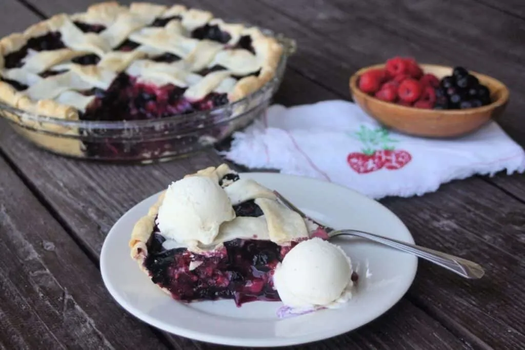 A slice of berry pie on a plate with scoops of vanilla ice cream and a fork. In the background is the remaining pie and a wooden bowl full of fresh berries. 