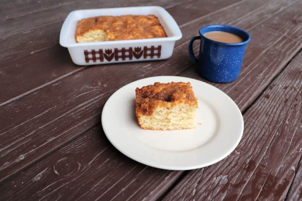 A piece of applesauce coffee cake on a white plate. A blue tin cup of coffee sits behind it, the remaining cake in square pan in the background.