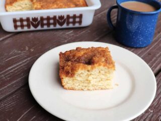 A piece of applesauce coffee cake on a white plate. A blue tin cup of coffee sits behind it, the remaining cake in square pan in the background.
