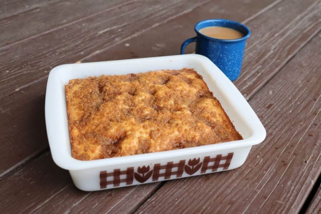 A streusel topped coffee cake in a white square pan with a blue tin cup of coffee behind it.