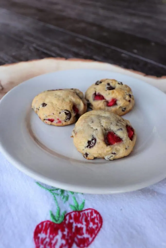 3 cookies on a plate that is sitting on a napkin with strawberries embroidered on it.