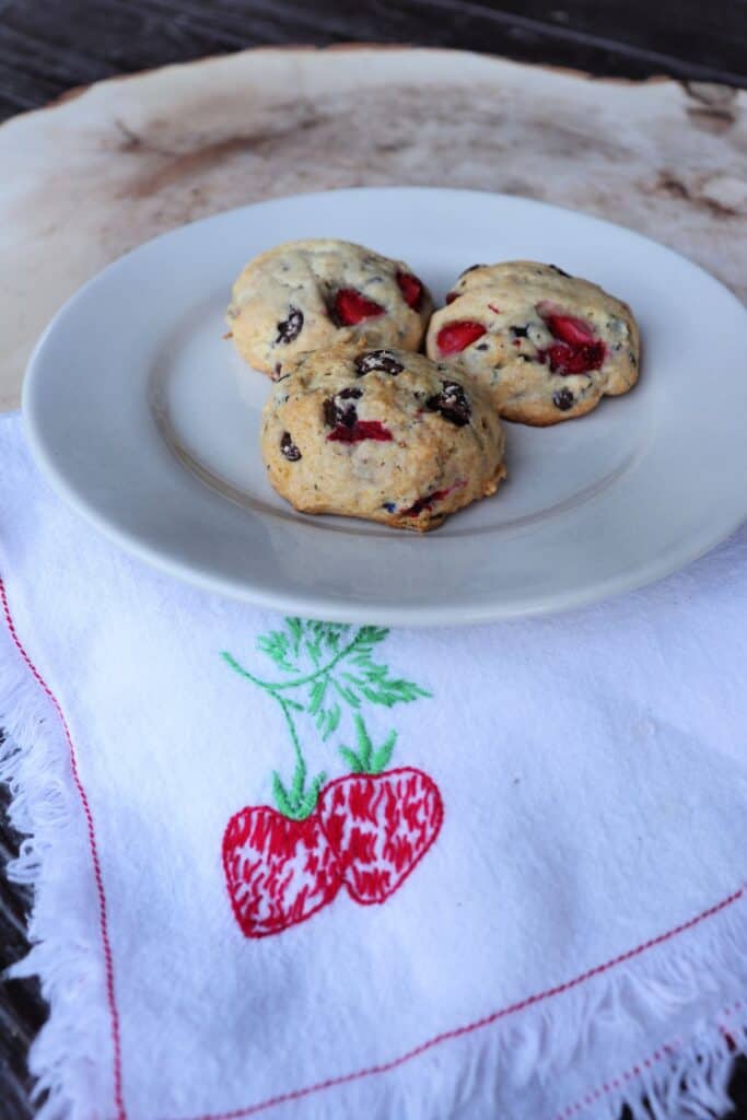 3 cookies with chocolate chips on a plate that is sitting on a napkin with strawberries embroidered on it.