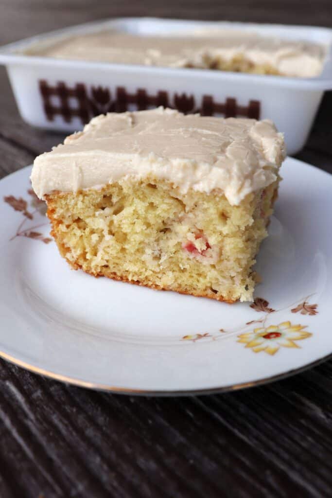 A Close up of piece of cake showing bits of rhubarb on a plate. Remaining cake still in pan sits in the background.