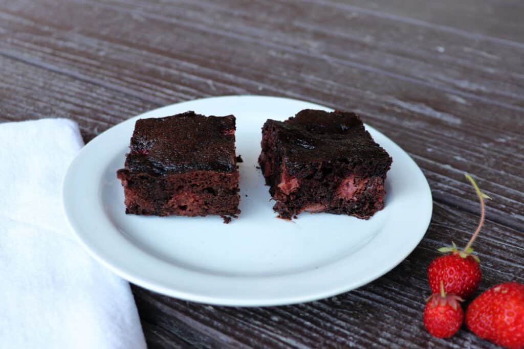 Two brownies on a plate exposing red bits of strawberries inside, a white napkin sits to the left and fresh strawberries sit to the right of the plate.