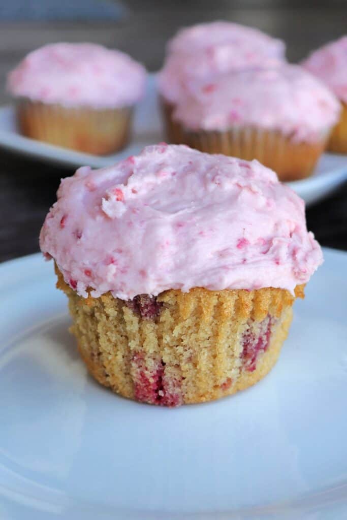 A raspberry cupcake with pink frosting sits on a white plate, additional cupcakes sit in the background.