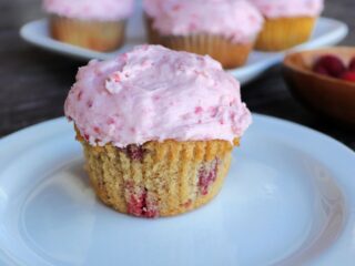 A raspberry cupcake with pink frosting sitting on a white plate. A platter of more cupcakes in the background.