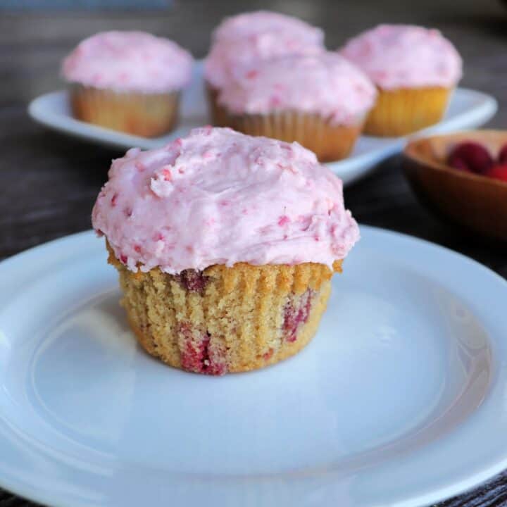 A raspberry cupcake with pink frosting sitting on a white plate. A platter of more cupcakes in the background.