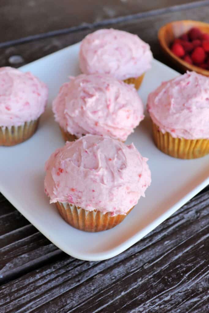5 raspberry cupcakes with pink frosting sit on a square white plate. A wooden bowl full of red raspberries sits in the background.