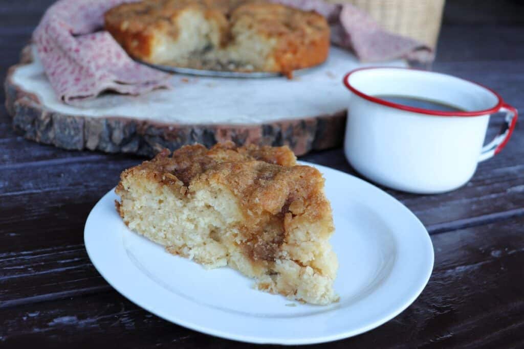 A slice of apple coffee cakes sits on a white plate. A red and white mug full of black coffee next to it. The remaining coffee cake sits on a board in the background.