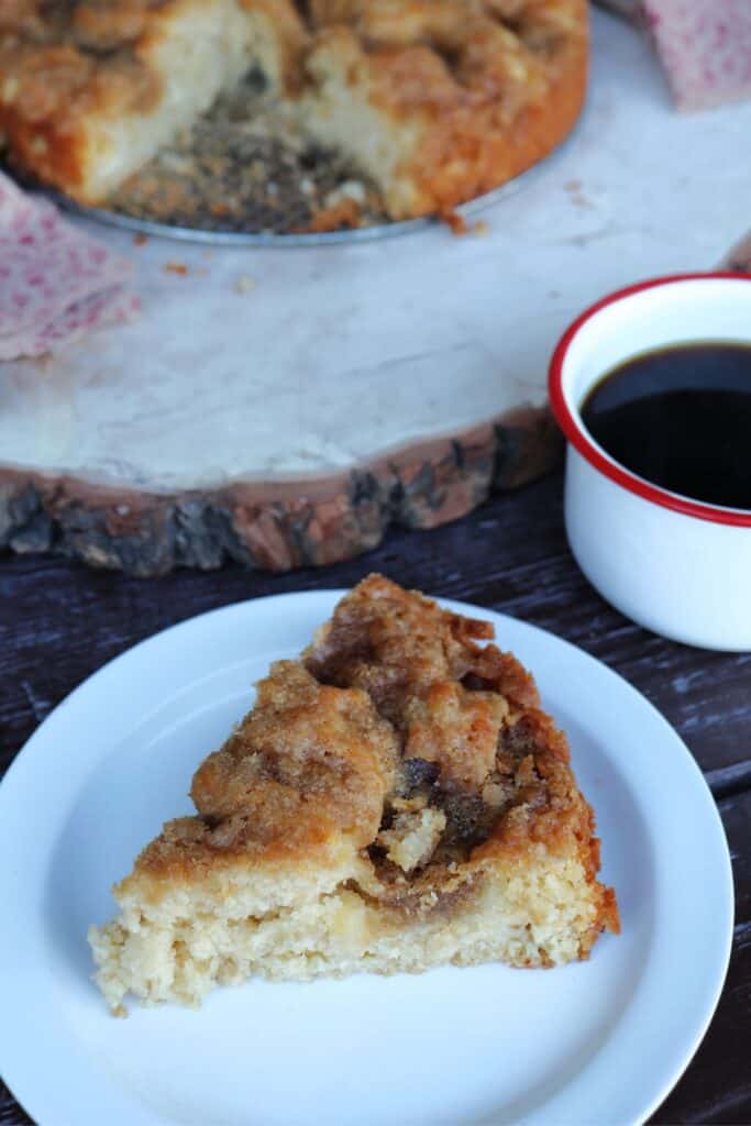 A slice of apple coffee cake sits on a white plate with a red and white mug full off coffee behind it. 