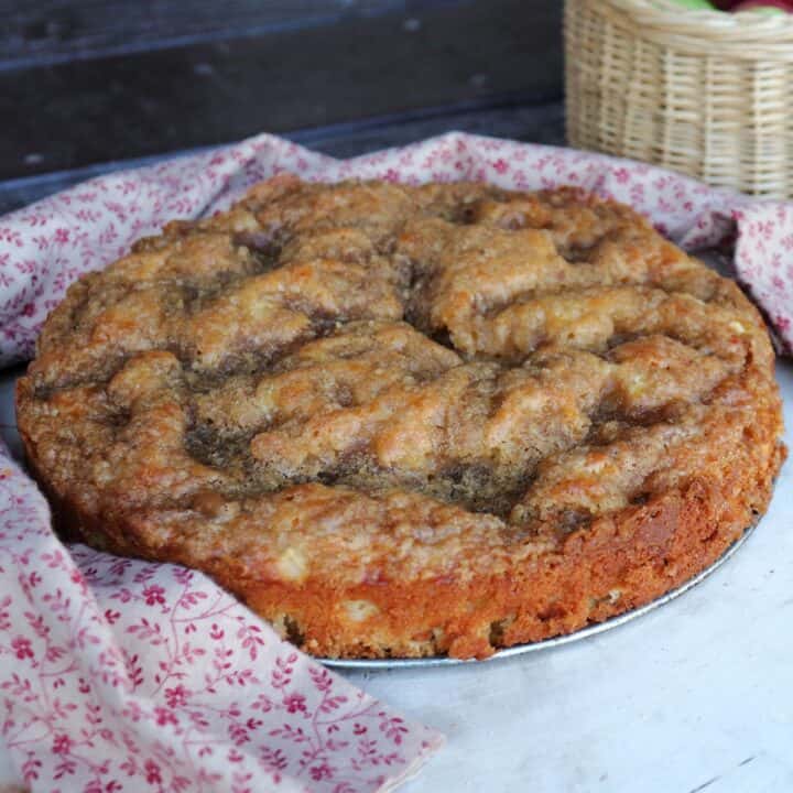 A round apple coffee cake sitting on a board surrounded by a red cloth.