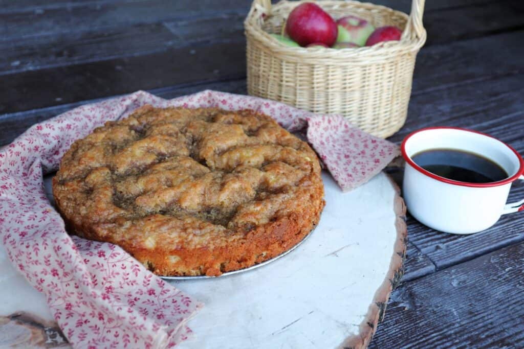 A round apple coffee cake sits on a board surrounded by a red flowered cloth. Behind the board is a basket full of red and green apples. A white and red cup full of black coffee sits tot he right.