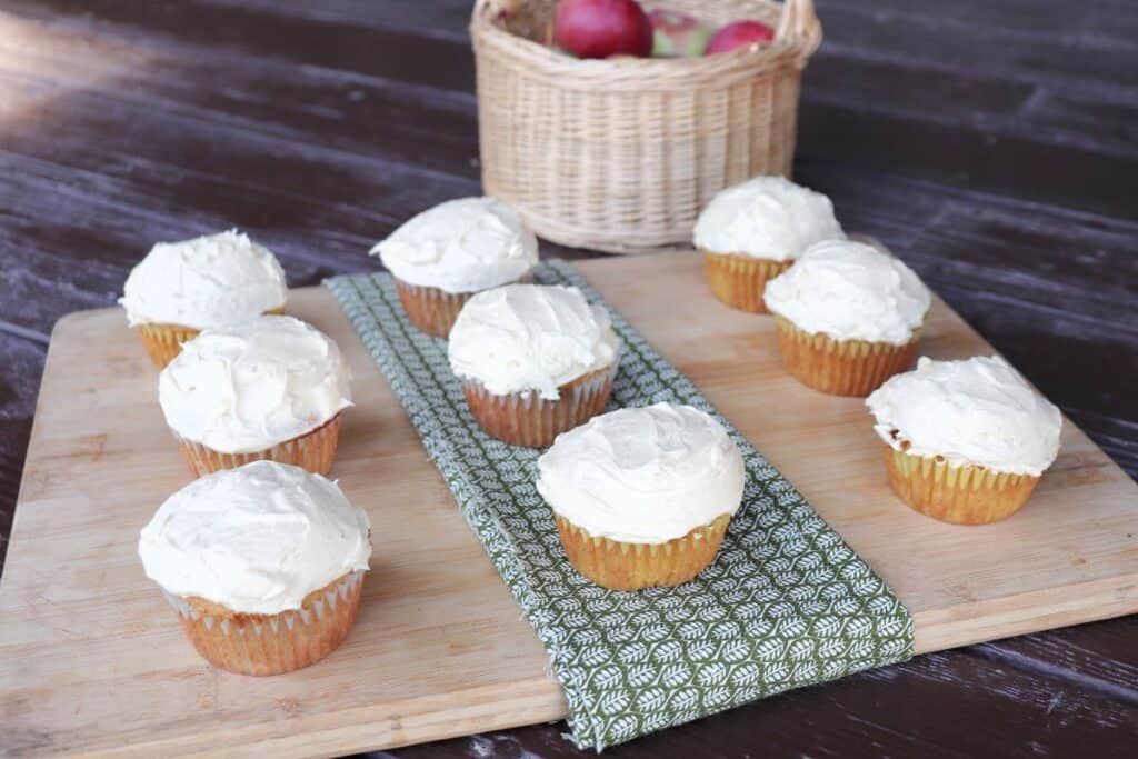 Apple cider cupcakes sit on a board with a basket of fresh apples in the background.
