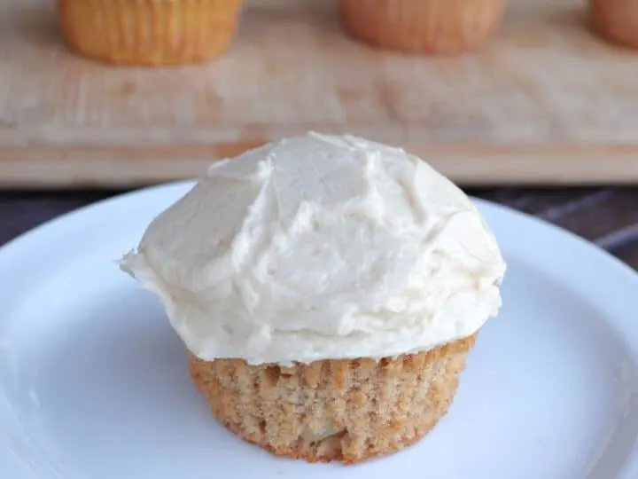 A frosted apple cider cupcake sitting on a white plate, more cupcakes in the background.