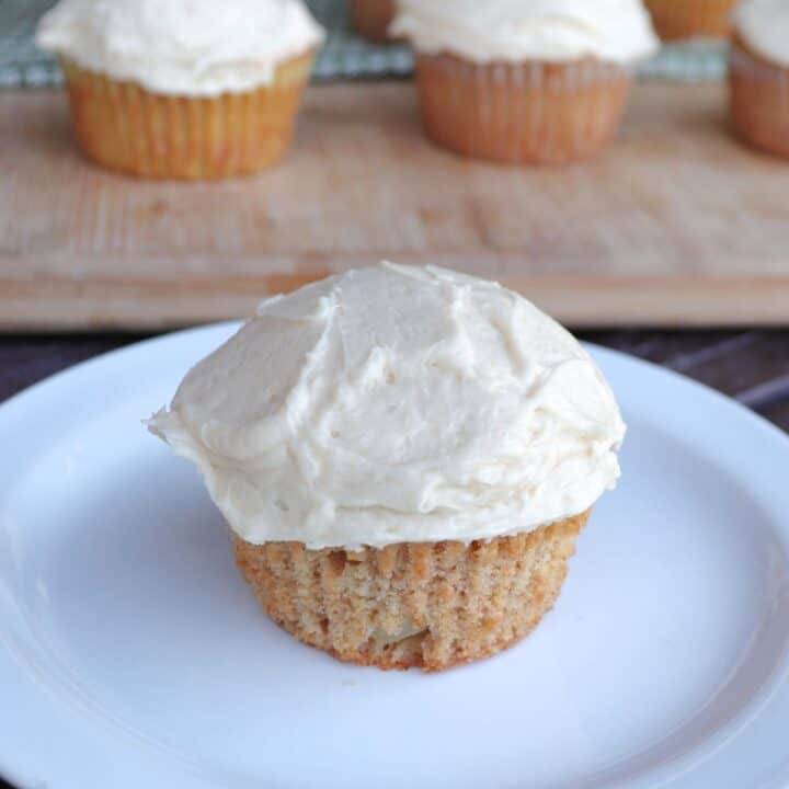 A frosted apple cider cupcake sitting on a white plate, more cupcakes in the background.