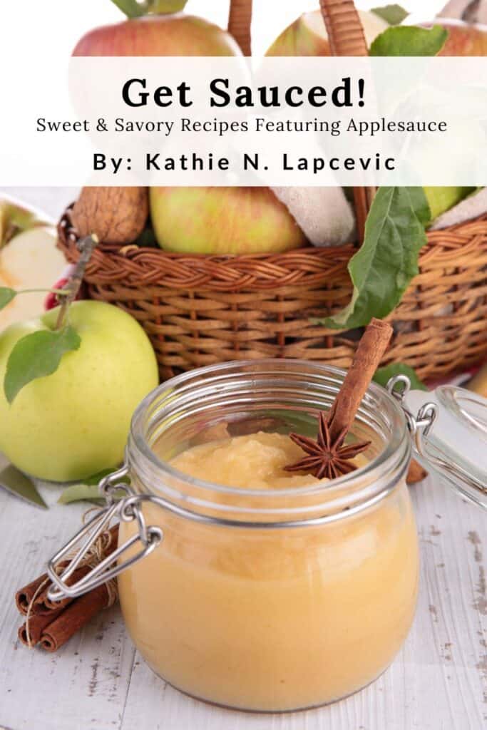 An open jar of applesauce with cinnamon stick sticking out of it. A basket of fresh apples in the background. Text overlay reads: Get Sauced! Sweet & Savory Recipes Featuring Applesauce by Kathie N. Lapcevic.