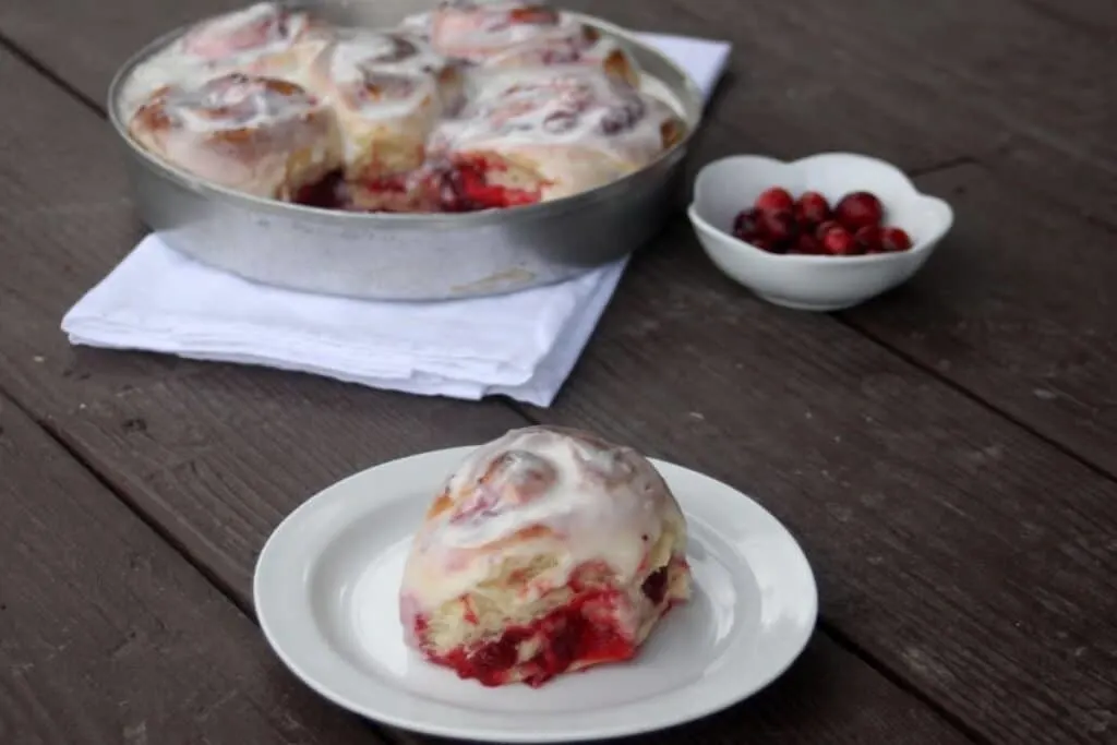 A frosted cranberry sweet roll sits on a small plate. A cake pan with more rolls sits in the background with a small bowl of fresh cranberries next to it.
