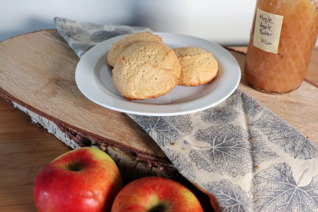 Cookies on a plate with fresh apples sitting in front of it and a jar of apple butter in the background.