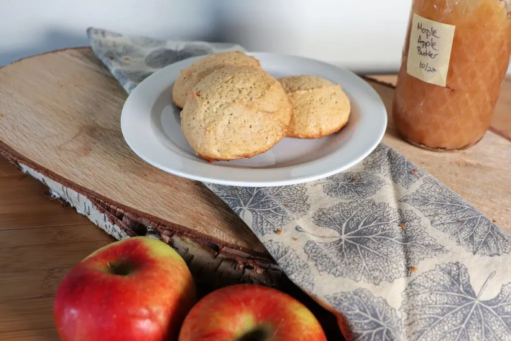 Cookies on a plate with fresh apples sitting in front of it and a jar of apple butter in the background.