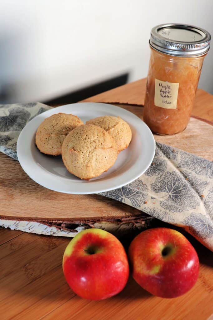 Cookies on a plate with fresh apples sitting in front of it and a jar of apple butter in the background.