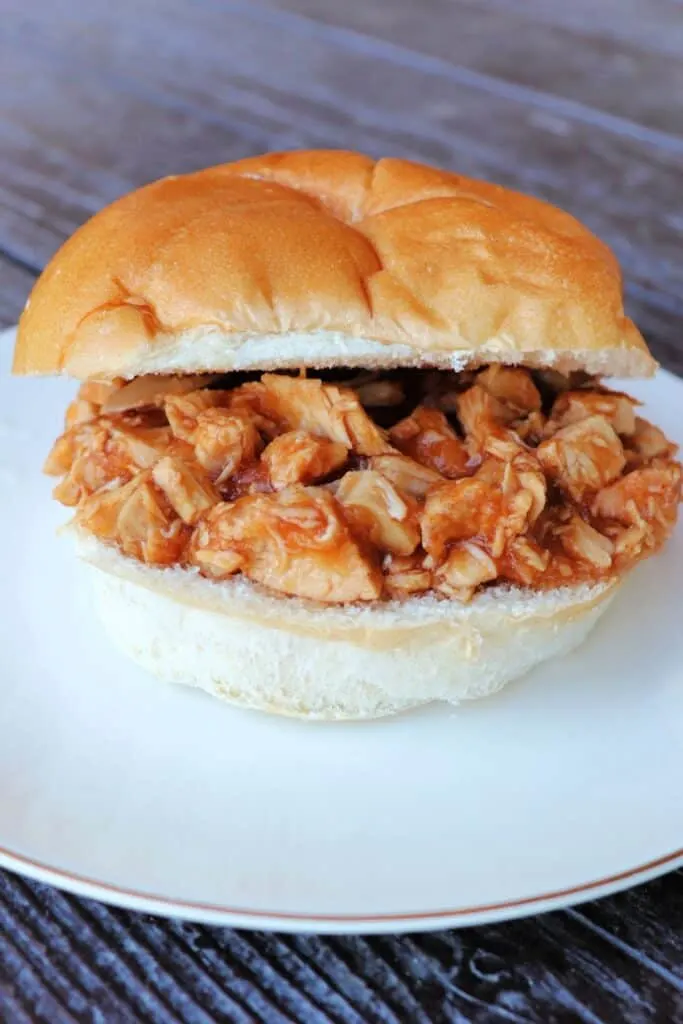 A bun that has been sliced open and filled with applesauce bbq chicken sits on a white plate.