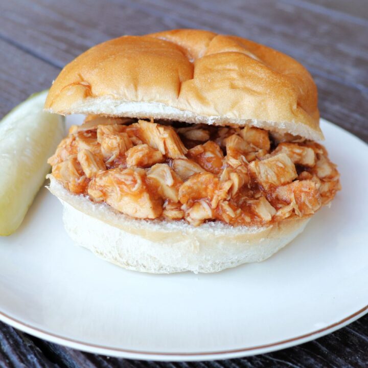 A split bun sitting on a plate with applesauce bbq chicken sitting in between the slices. A pickle spear sits on the plate.