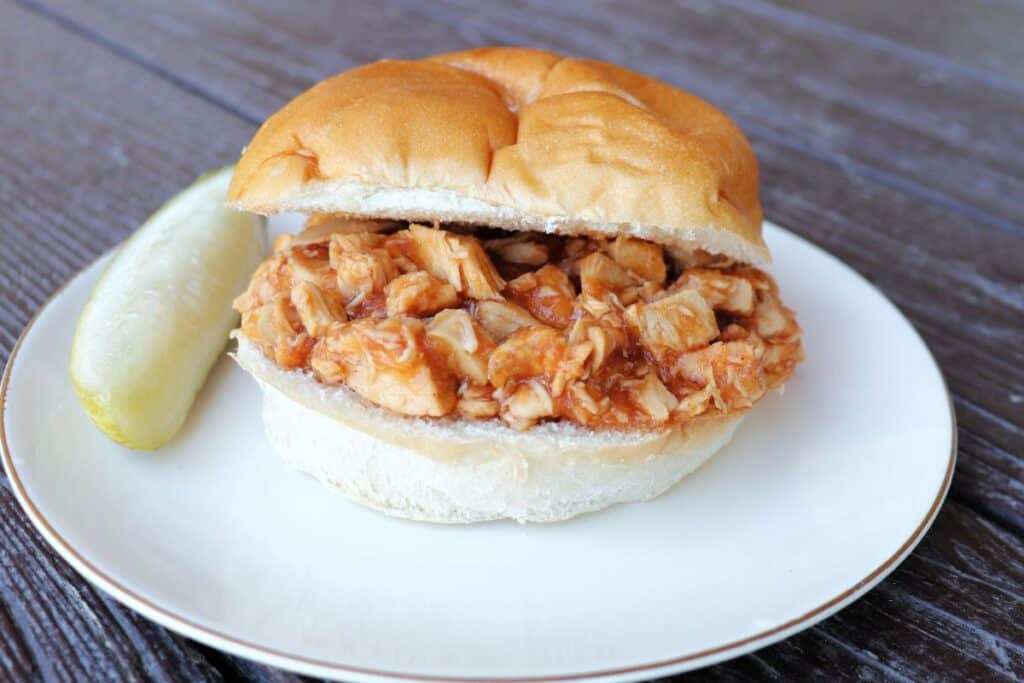 A split bun sitting on a plate with applesauce bbq chicken sitting in between the slices. A pickle spear sits on the plate.