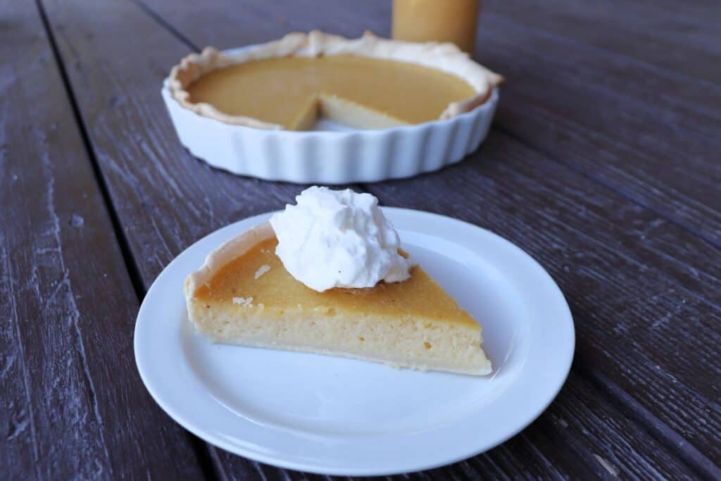 A slice of applesauce pie as seen from the side with a dollop of whipped cream on top, the remaining pie sits in the background. 