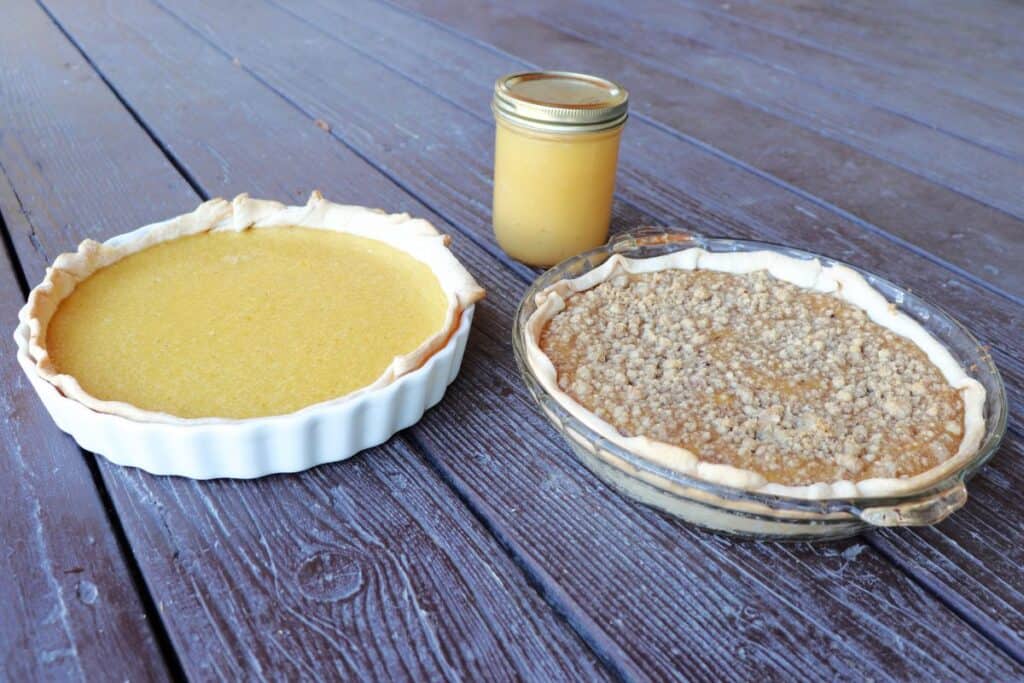 Two applesauce pies, one without a topping in a white pie plate, the other with a crumble topping in a clear glass pie plate. A glass jar full of applesauce sits behind them.