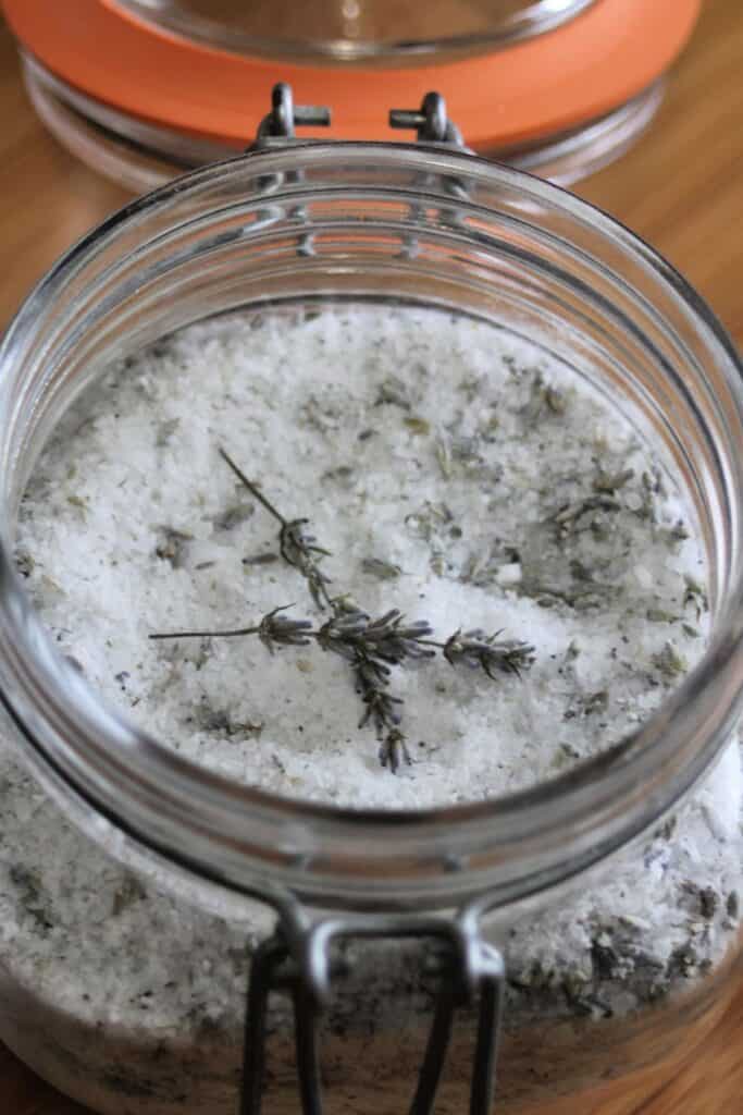An open jar of bath salts with dried lavender stems sitting on top. 