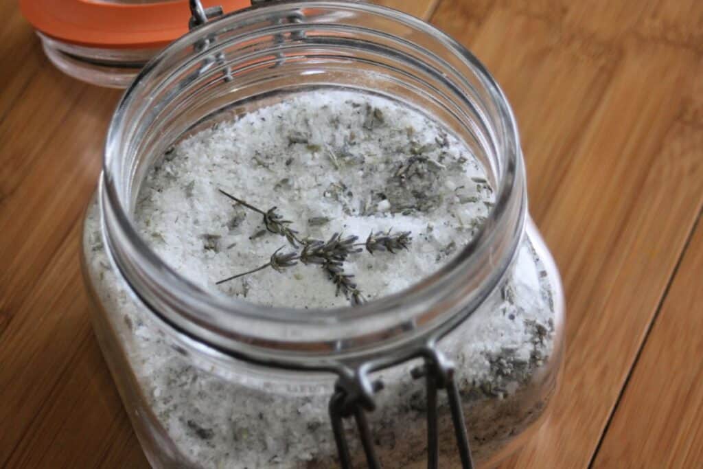 An open jar of bath salts with dried lavender stems sitting on top. 