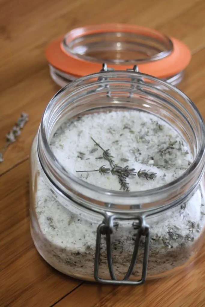 An open jar of bath salts with dried lavender stems sitting on top and a dried stem of lavender sitting next to it.