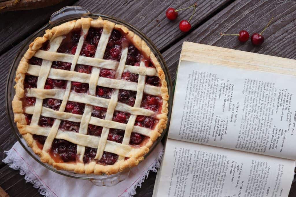 A lattice topped cherry pie sitting next to an open book.