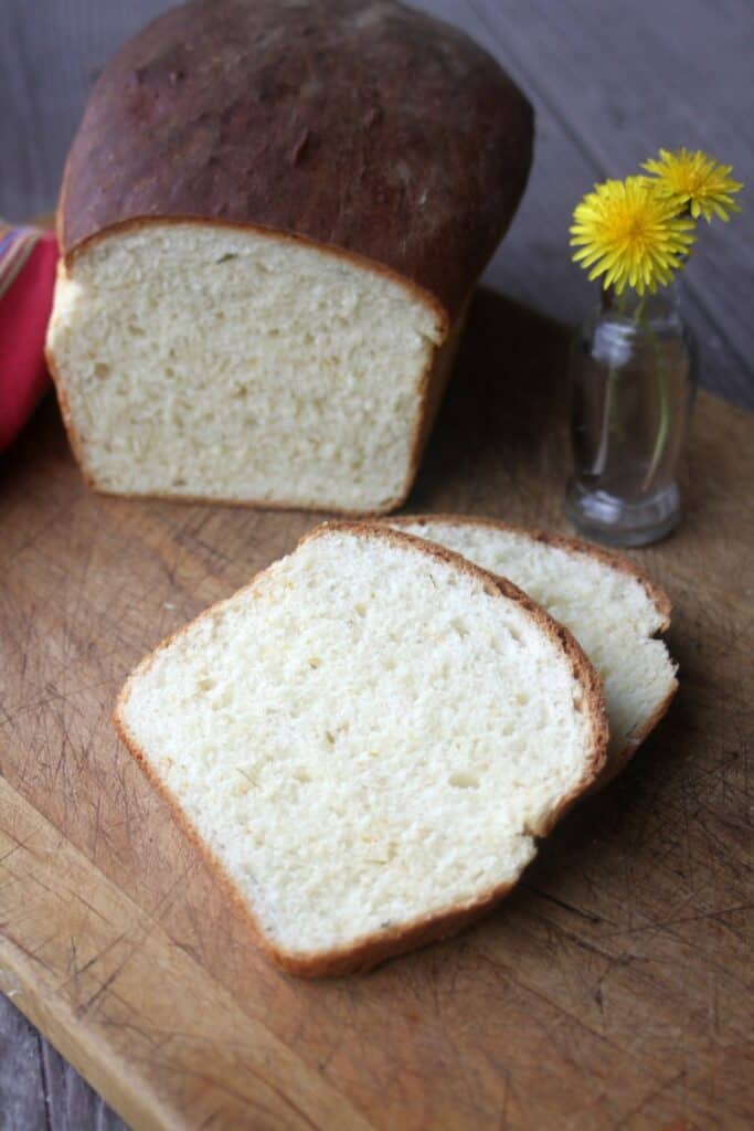 Slices of bread on a cutting board with the remaining loaf in the background. A small vase full of dandelion flowers sits on the board.