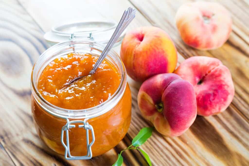 An open jar of peach jam with a spoon sticking out of it and fresh peaches on the table next to the jar.