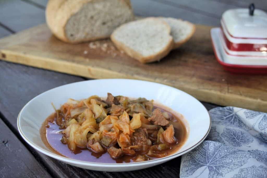 A bowl of cabbage and beef soup sits on a table. A napkin sits to the right of the bowl.  A cutting board sits in the background with a loaf of bread and butter dish on top.