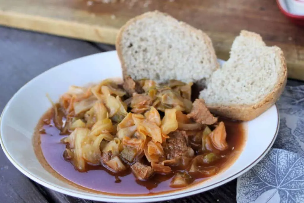 A bowl of cabbage and beef soup sits on a table, 2 slices of bread sit on the edge of the bowl.