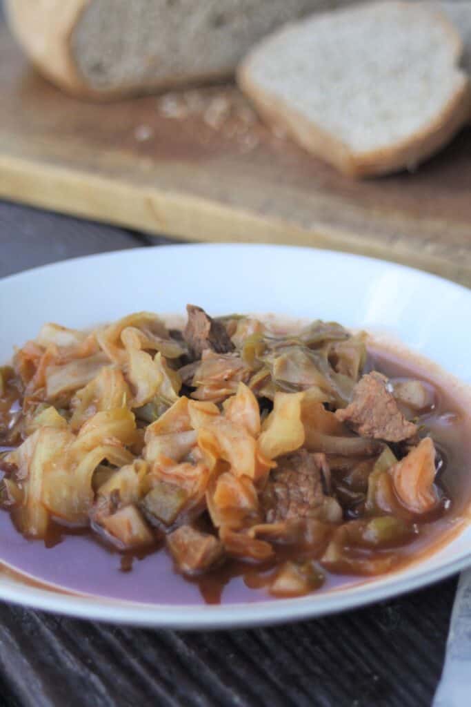 A bowl of beef and cabbage stew sits on a table. A cutting board with slices of bread sits in the background.