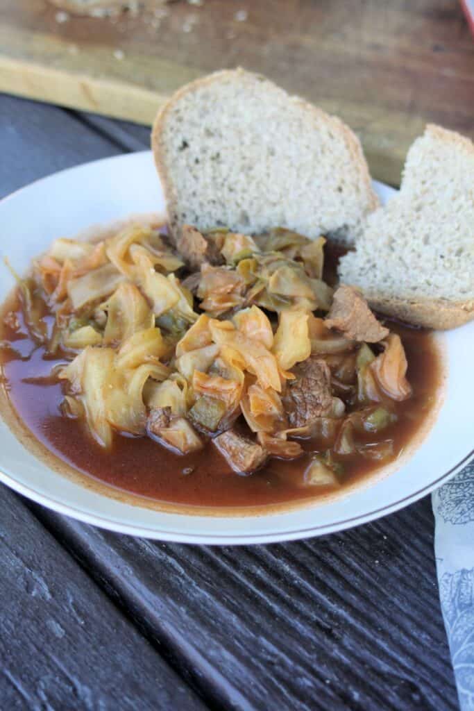 A bowl of cabbage and beef soup sits on a table, 2 slices of bread sit on the edge of the bowl.