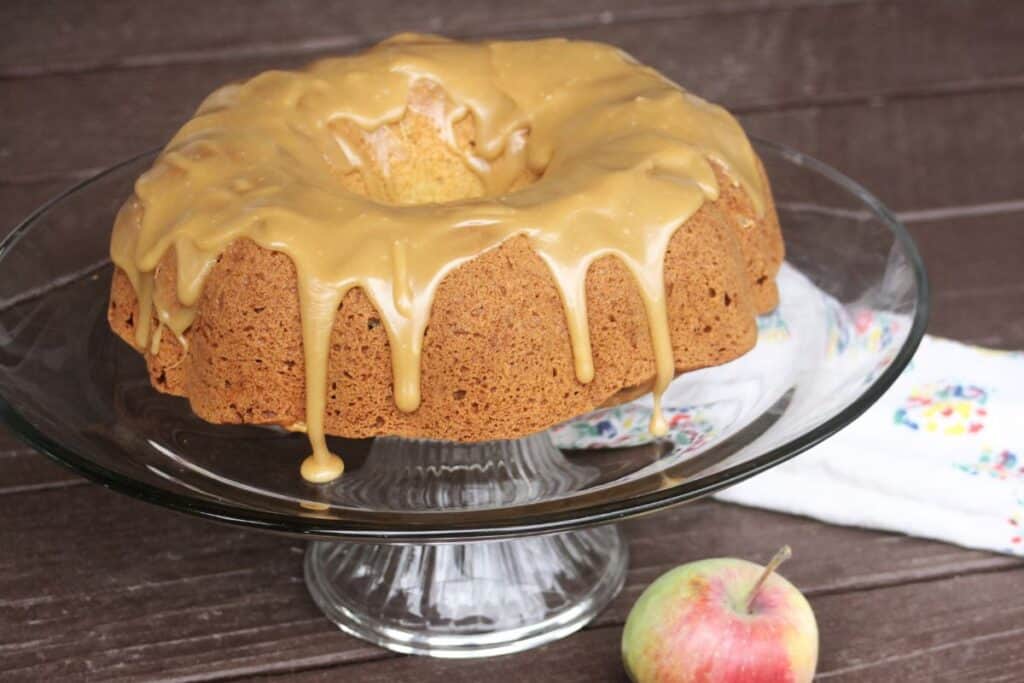 A caramel glazed apple bundt cake sits on a glass cake plate. A fresh apple and cloth napkin sit on the table. 