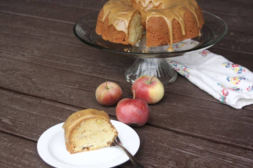 A slice of frosted cake on a plate with a fork. The remaining cakes sits on a cake plate in the background, fresh apples on the table.