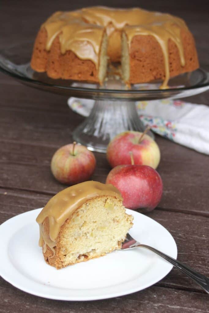A slice of frosted cake on a plate with a fork. The remaining cakes sits on a cake plate in the background, fresh apples on the table.