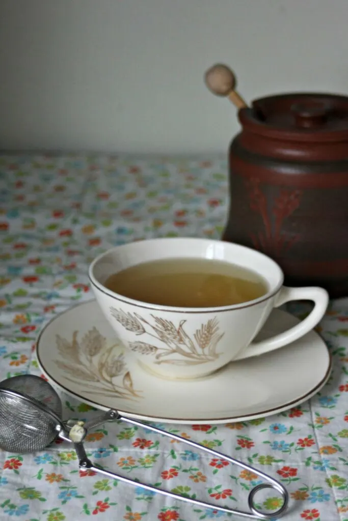 A cup of herbal tea sits on a table covered in a floral cloth - a jar of honey sits in the background. 