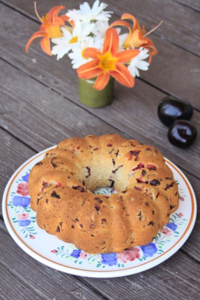 A bundt cake sits on a floral cake plate. A vase of daisies and lilies sit in the background.