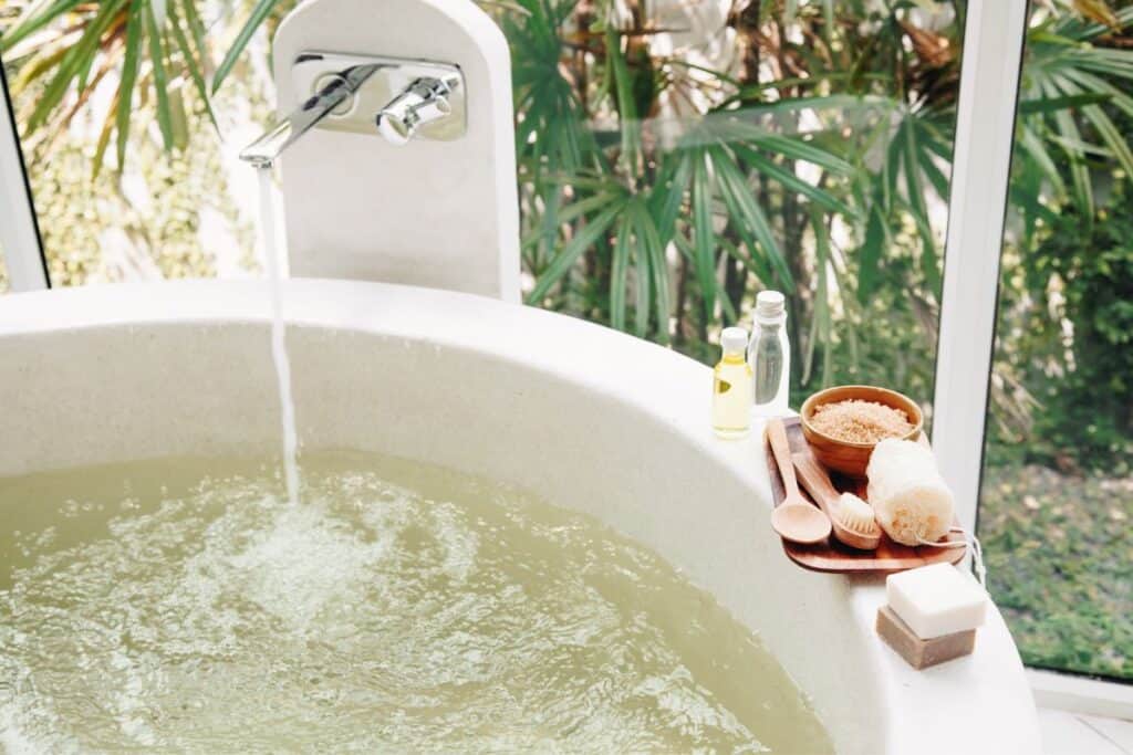 Running water from a faucet filling up a bath tub. Soap, bottles of oil, and sponges sit on the rim of the tub.
