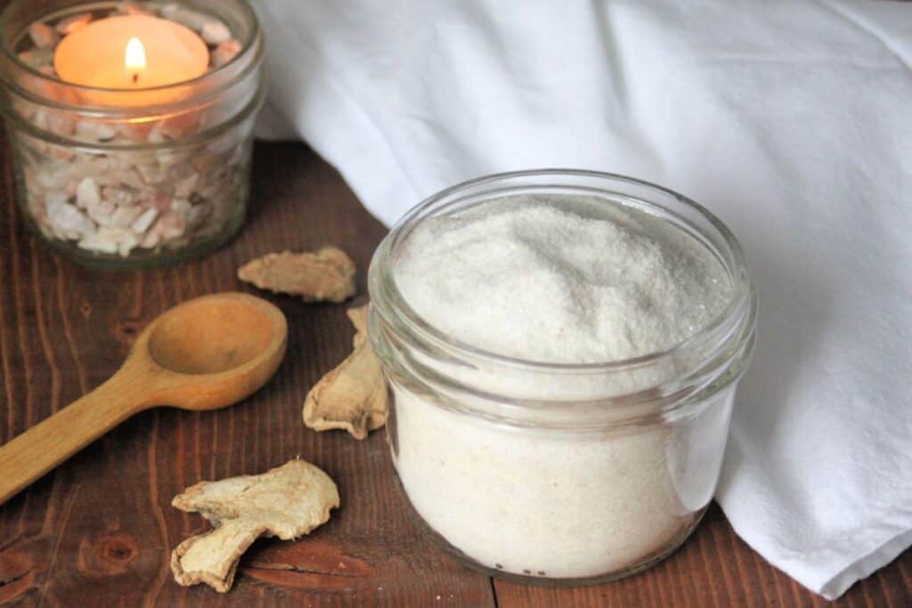 An open jar of ginger bath salts surrounded by a white cloth, wood spoon, slices of dried ginger, and a lit candle.